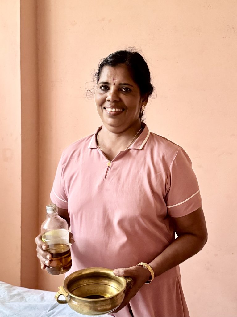Sarojam, a Female Therapist at Prakruthi Ayurveda Cherai Beach, holds a bottle of Ayurvedic oil and a brass vessel for massage.